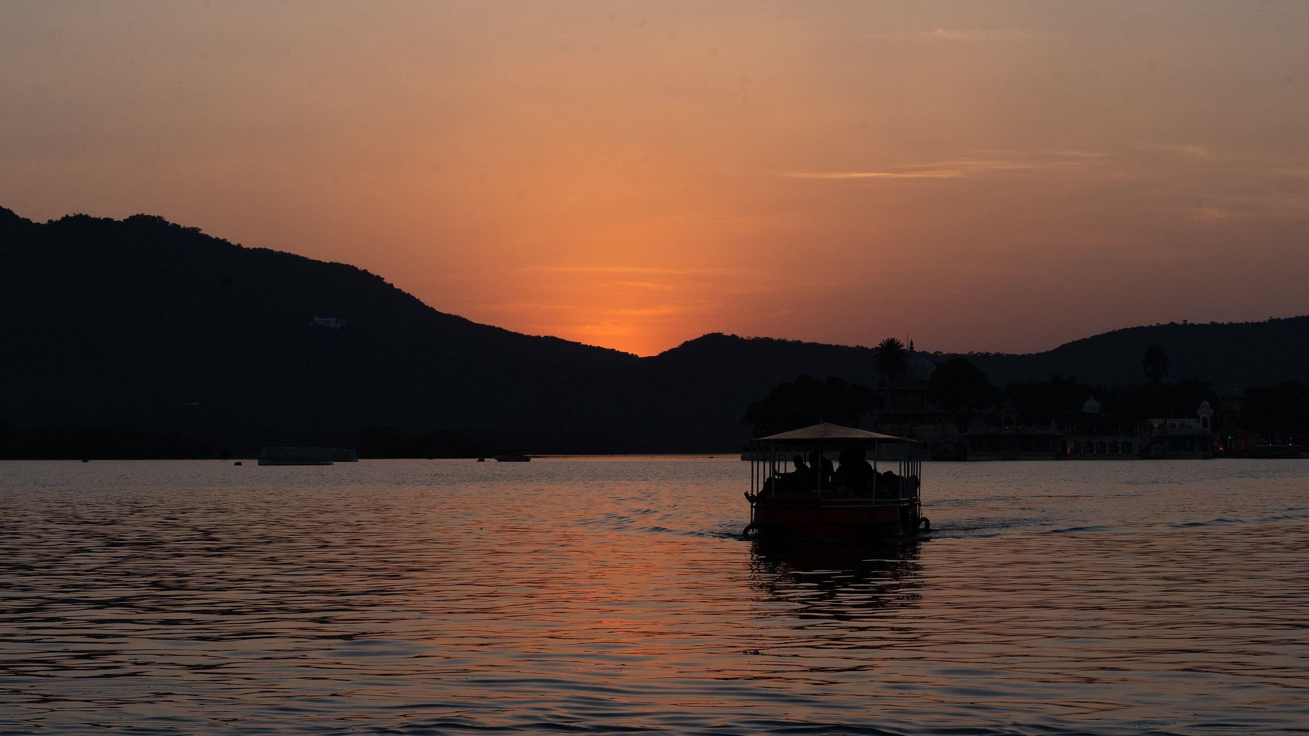 Sunset over Lake Pichola in Udaipur: dark Aravalli hills against an orange sky and a covered boat on the water.