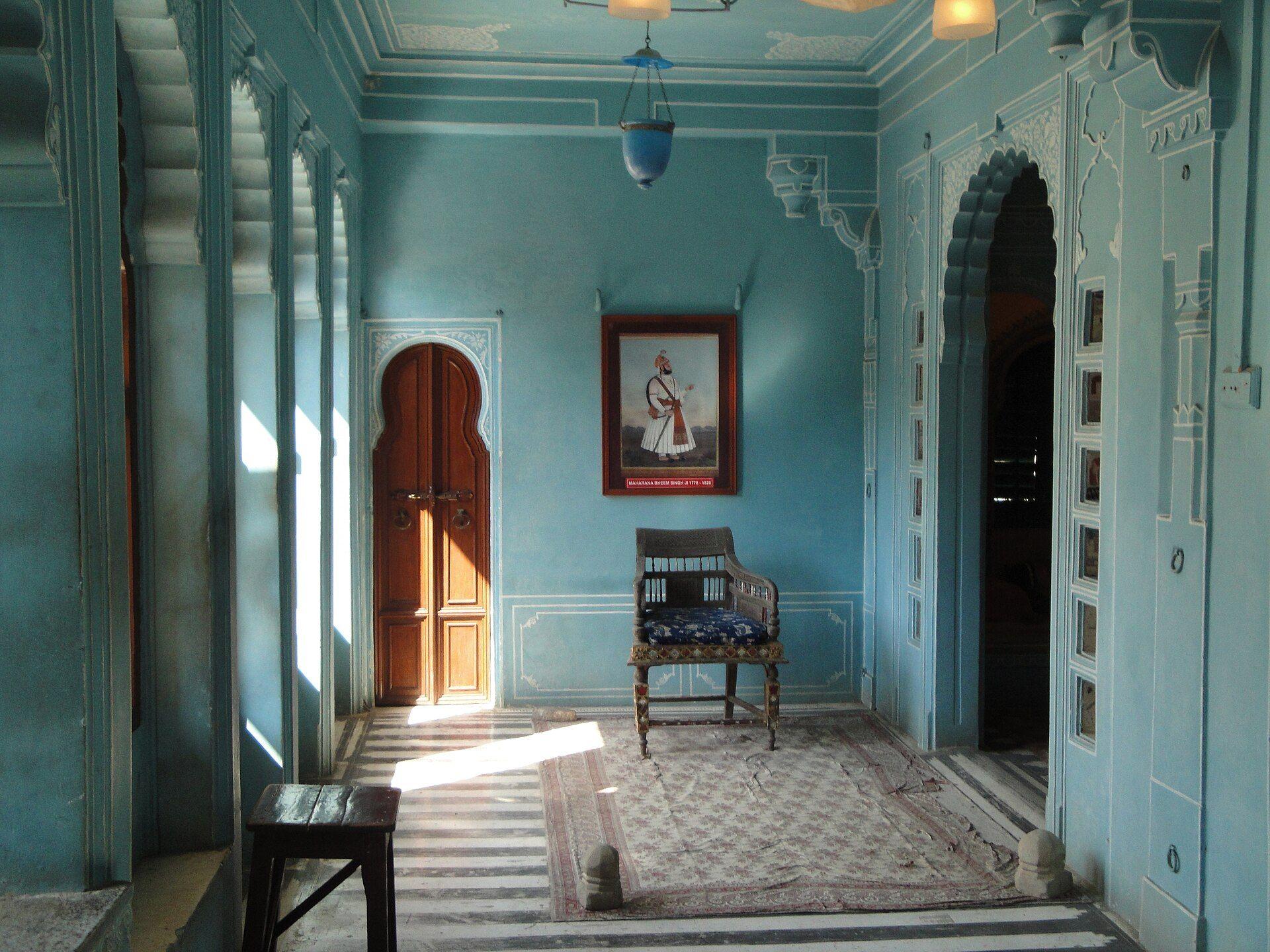 A quiet room in the City Palace Museum with pale blue walls, an arched wooden door, a chair and a portrait.