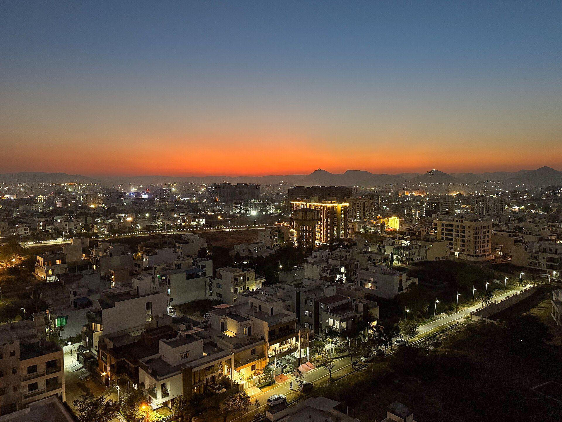 Udaipur's rooftops at dusk, with lit windows and streets under an orange horizon.
