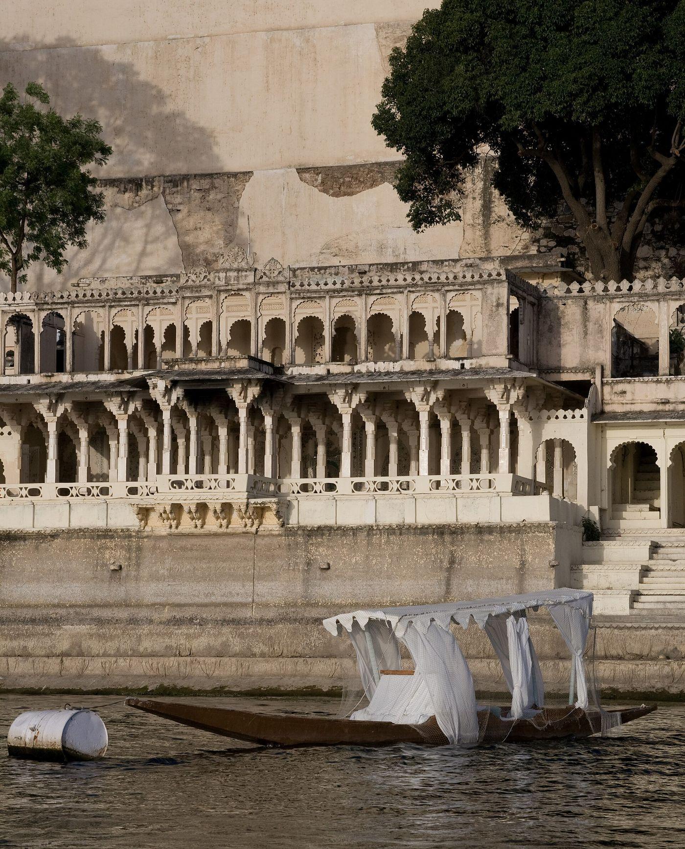 A small boat with a white canopy moored below an arcaded building on the shore of Lake Pichola.