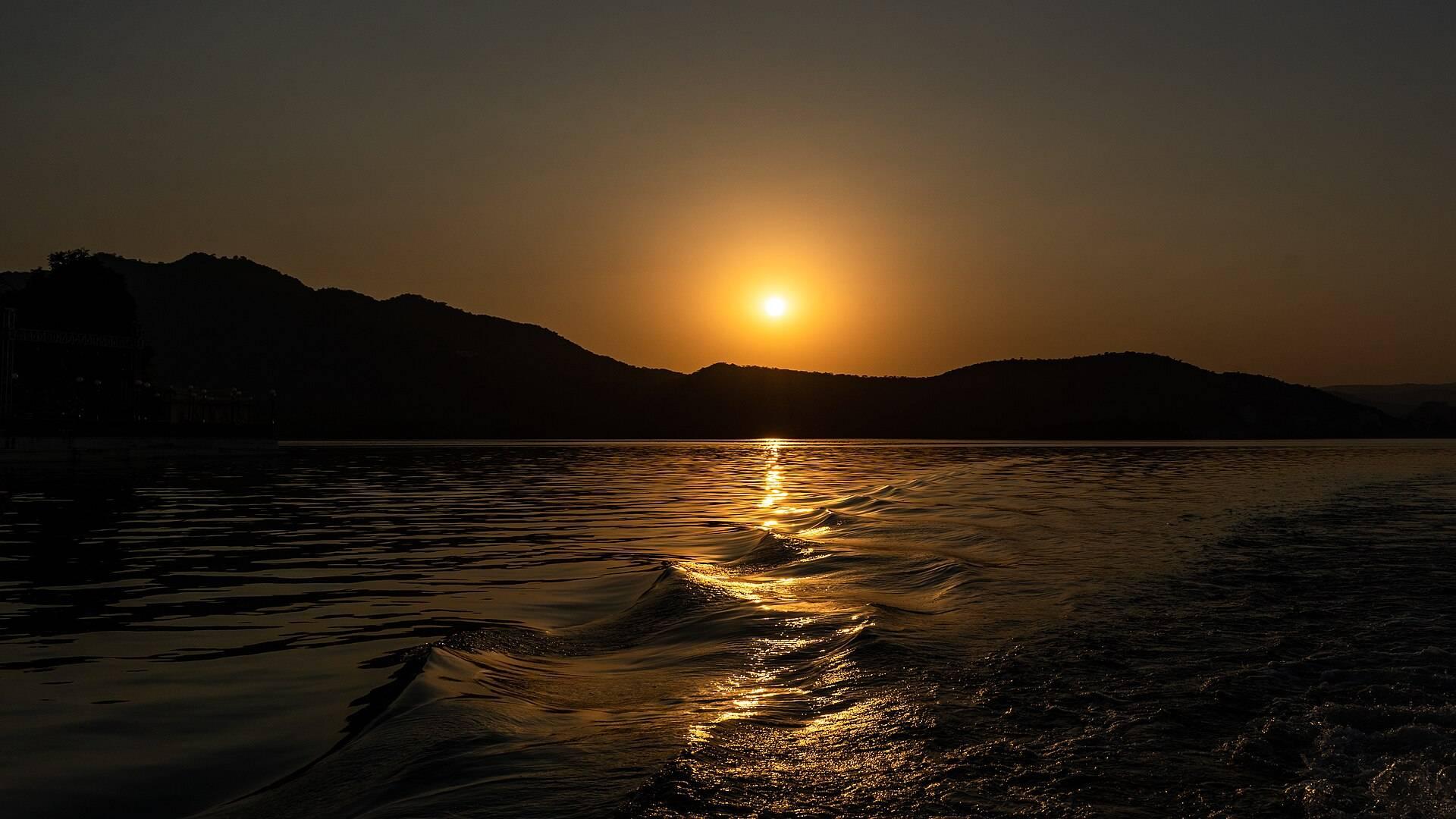 The setting sun behind dark hills, its gold light running across the water of Lake Pichola.